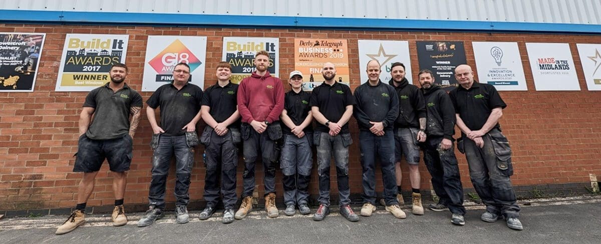 Image for the Gowercroft Assembly team standing in front of the factory in front of a wall where Gowercroft's awards are displayed. They are all wearing polo shirts, tradesman style combat trousers and steel toe cap shoes. The team leader wears a maroon sweatshirt.