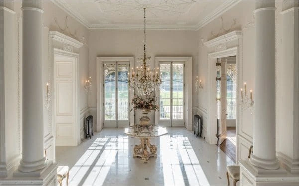 Bright, elegant hallway with tall white columns, decorative plasterwork, chandeliers and full-height glazed doors leading to the garden.