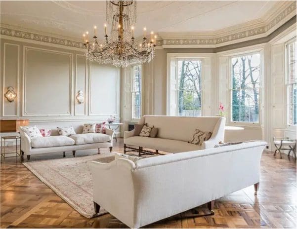 Elegant period living room with tall timber windows, decorative wall panelling, a crystal chandelier, parquet flooring and cream sofas.