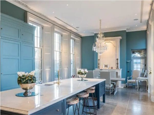 Elegant teal-blue kitchen and dining room with tall white timber windows, a large marble island, crystal chandelier and period fireplace.