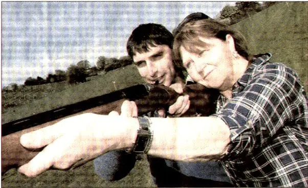 A newspaper clipping photo showing Gowercroft Director David Brown assisting a woman as she aims a shotgun during a charity clay pigeon shoot. He is demonstrating proper technique in a countryside setting, with grassy hills in the background.