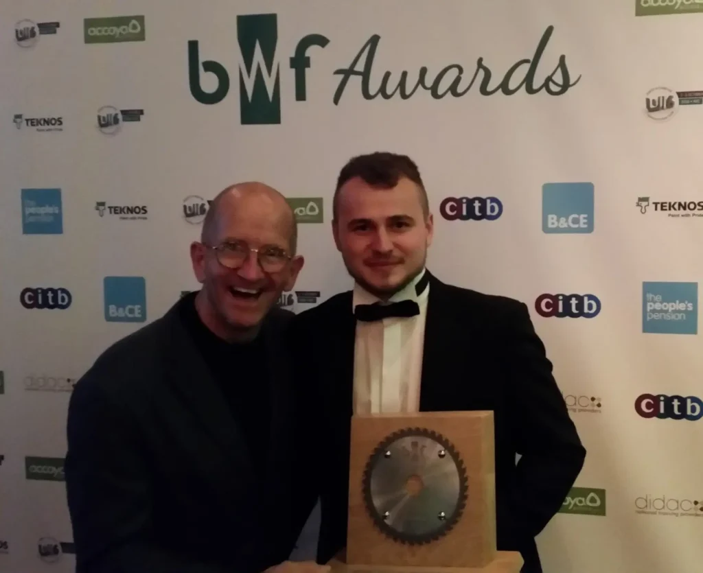 Greg Kasowicz holding his award at the 2016 BWF Awards ceremony, standing beside Eddie 'the Eagle' Edwards in front of the official event backdrop featuring sponsor logos.
