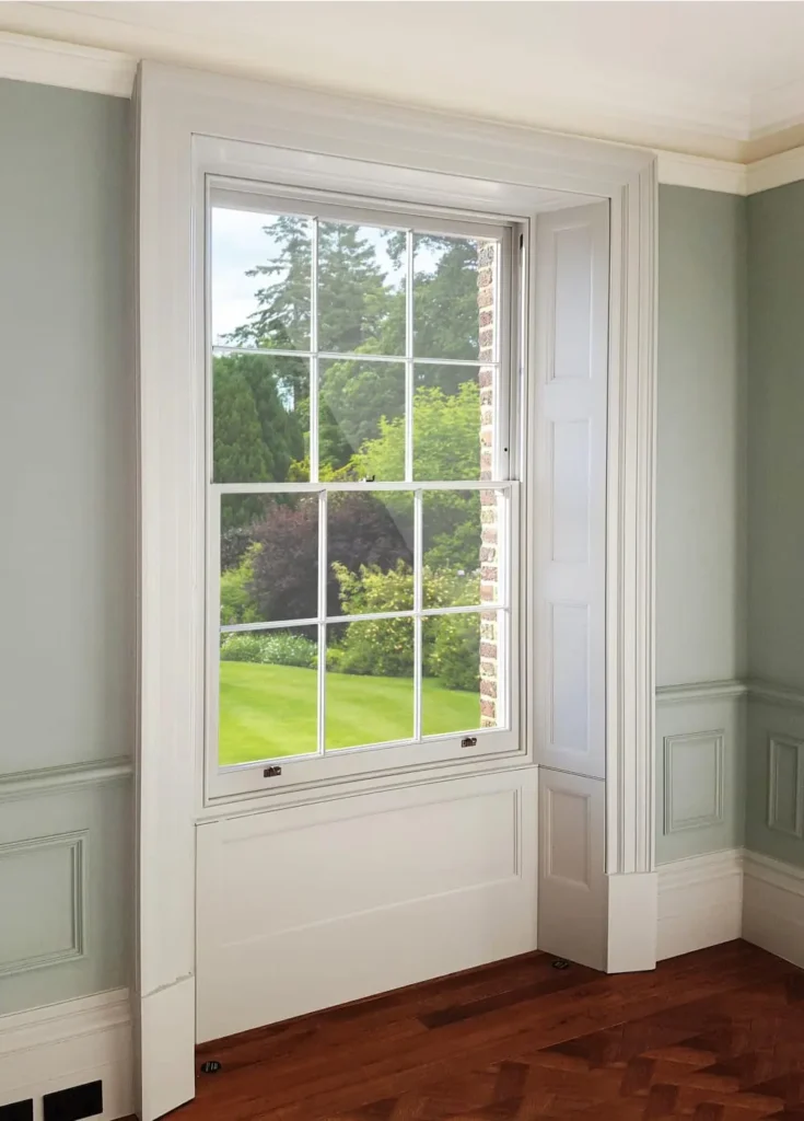 Interior view of a Winston heritage sliding sash window by Gowercroft Joinery, featuring slim timber glazing bars and LandVac vacuum glass, set in a traditionally styled sash box with decorative paneling and white microporous finish. Lush green garden visible through the clear, reflection-free glass.