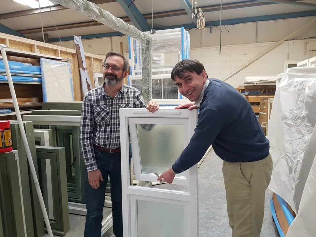Gowercroft founders and directors Chris and David in the workshop, smiling while demonstrating one of their bespoke timber windows as part of the company’s 20-year anniversary.