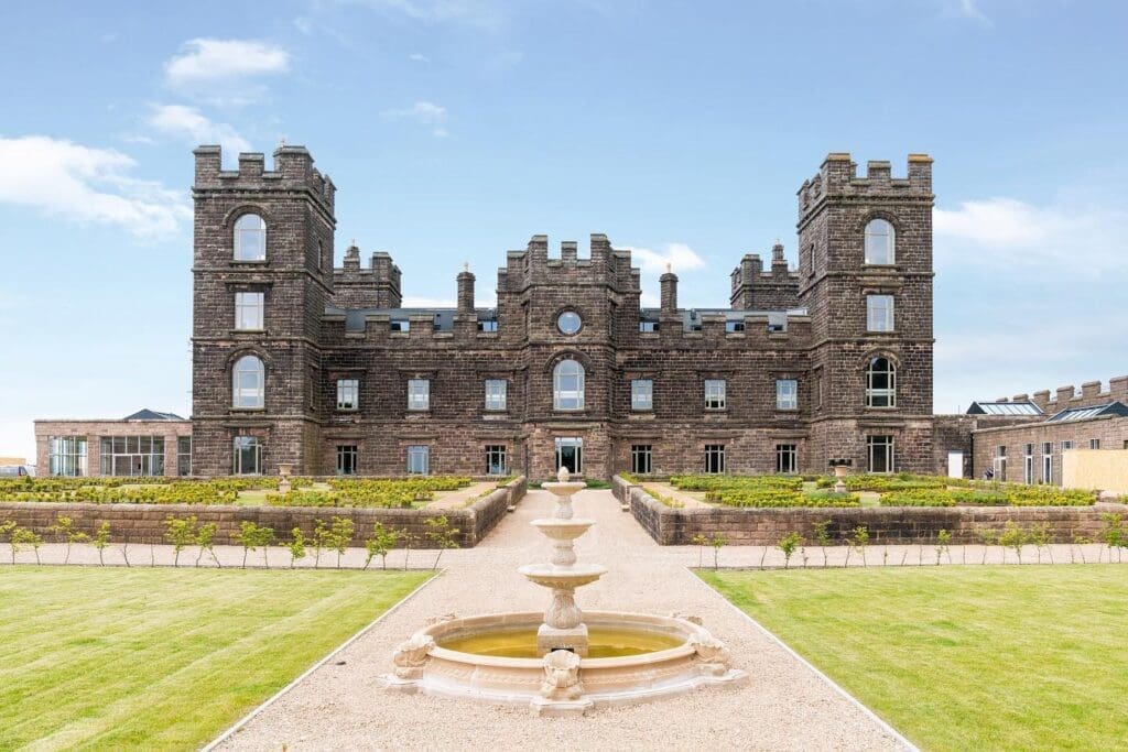 Restored exterior of Riber Castle with landscaped grounds
A full-frontal view of Riber Castle in Matlock, Derbyshire, showing the dark stone façade, castellated towers, and symmetrical layout. The formal gardens feature topiary, a circular fountain in the foreground, and gravel pathways. All visible windows are custom-made timber casements from Gowercroft. Image courtesy of Dales and Peaks.