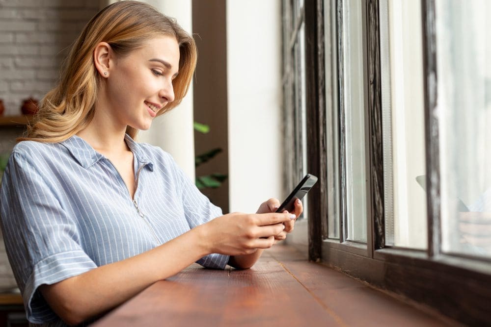 A young woman smiling and using her phone while standing beside a traditional timber sash window, with natural daylight illuminating her face.