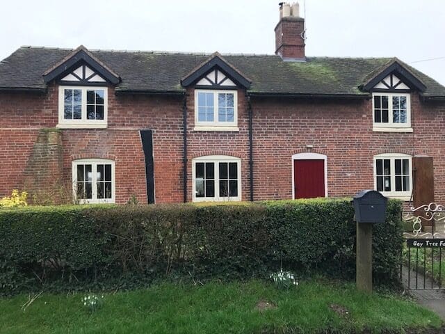A charming traditional red-brick cottage with timber casement windows and a red front door, set behind a neatly trimmed hedge and lawn. The classic cottage-style windows feature white-painted wooden frames and are perfectly suited to the character of the home, offering a visual example of how timber casement windows can complement period properties while delivering practical benefits.