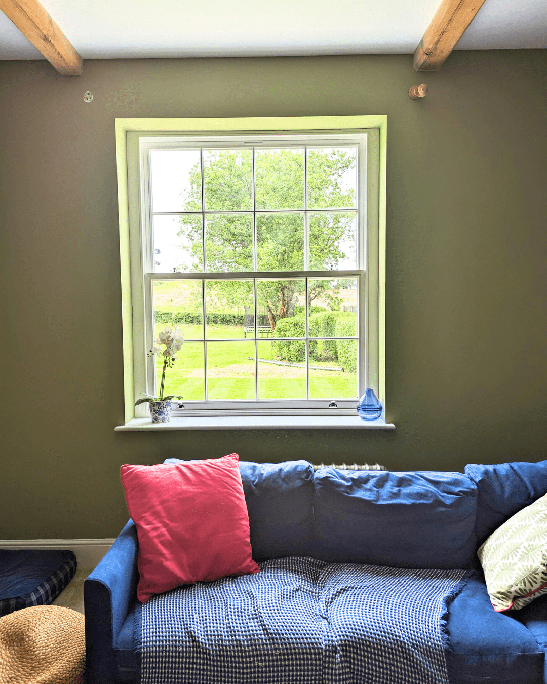 A cosy living room with a large white Winston Heritage Sliding Sash timber window by Gowercroft Joinery. The window provides a picturesque view of a lush green garden, enhancing the room's serene ambience. Inside, a comfortable blue sofa adorned with colourful cushions sits against the olive-green wall, underlining the room's warm and inviting atmosphere. The exposed wooden ceiling beams add a rustic charm, while the well-lit space highlights the window's elegant design and craftsmanship.