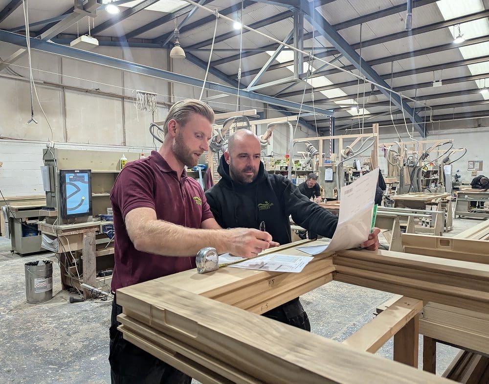 Two Gowercroft Joinery team members review paperwork together on top of stacked timber window frames in a busy workshop. The man on the left wears a burgundy polo shirt and points at the documents, while the man on the right wears a black hoodie and leans in to look. Machinery, timber components and workshop equipment fill the background.