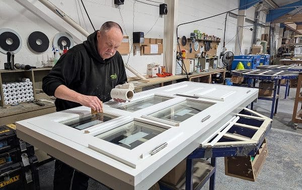 Tony from Gowercroft Joinery wet glazes a white timber door in the workshop, carefully applying glazing sealant around the glass panes. The door lies flat on a workbench, surrounded by tools, materials and workshop equipment.