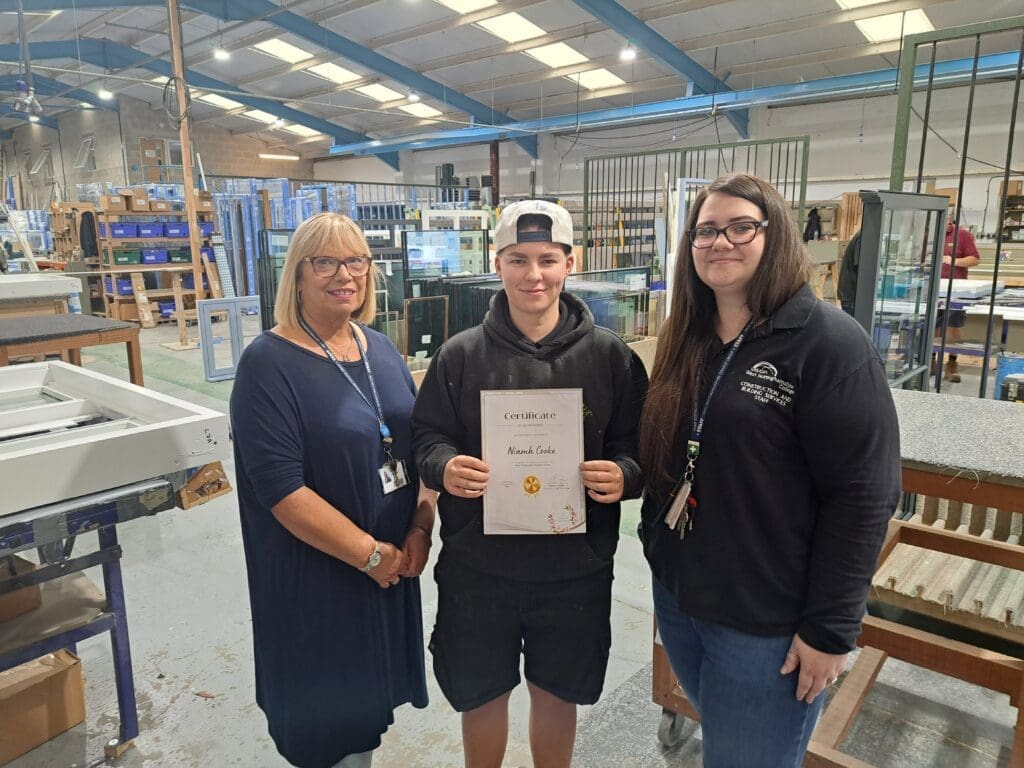 Apprentice Niamh Cooke holding her ‘Most Employable Student Award 2023’ certificate in the Gowercroft Joinery workshop, pictured with two college representatives