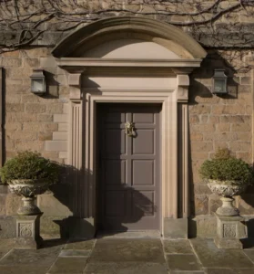 Traditional panelled front door set within an arched stone surround, flanked by wall lanterns and decorative planters outside a period stone building.