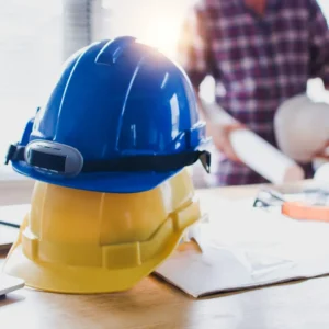 Blue and yellow hard hats on a desk with building plans, with a construction professional holding drawings in the background.