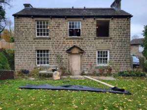 raditional stone house under renovation, with sash windows, an open upper window, a person visible inside, and garden materials laid across the lawn.