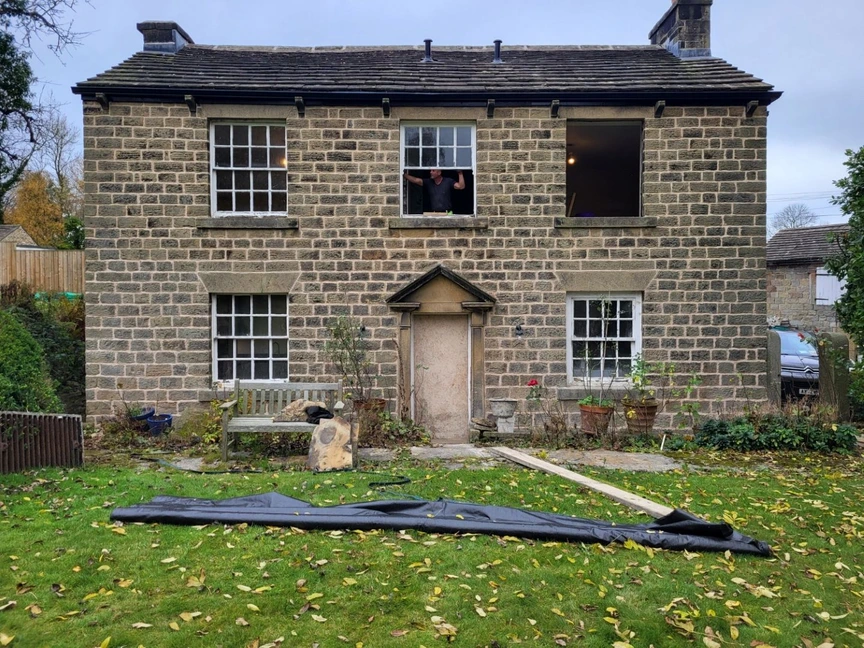 raditional stone house under renovation, with sash windows, an open upper window, a person visible inside, and garden materials laid across the lawn.