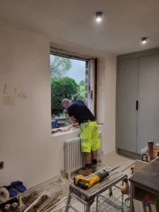 Worker removing an old window frame from inside a home renovation, with exposed masonry, tools, debris, and a garden visible through the opening.