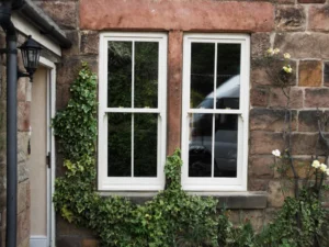 White timber sash windows set into a rustic stone wall, with climbing ivy, pale roses, and garden reflections in the glass.